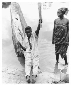 A boatman brings home a bat for soup to the banks of the Ubangi River in his dugout canoe. His wife wears a traditional print wrap dress.