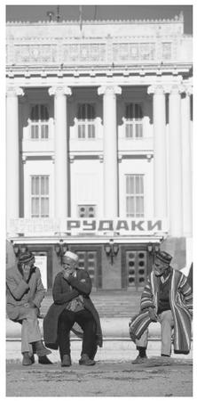 Men wearing traditional Tajikistani clothing sit in front of a Soviet–constructed theater.