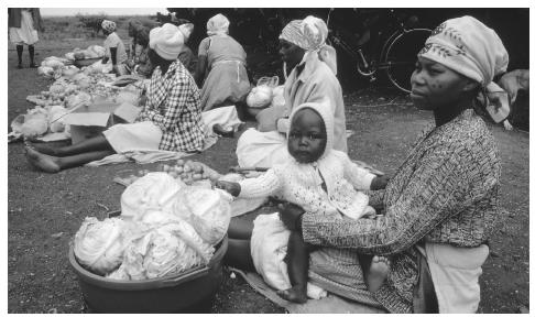 Women and children sit alongside a road with food. Women are responsible for the care of infants, and they typically carry their babies on their backs.