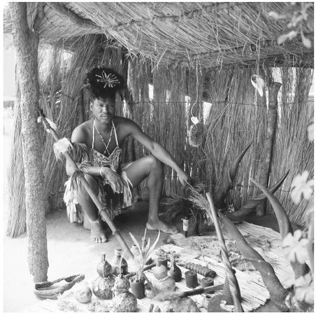 A healer from the Tonga, Zambia, sits in his grass hut. Various gourds, boxes, and other items are spread before him.